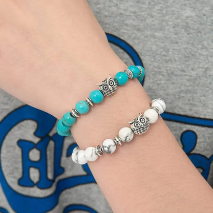 Hand wearing turquoise and white beaded bracelets with silver owl charms on gray shirt background